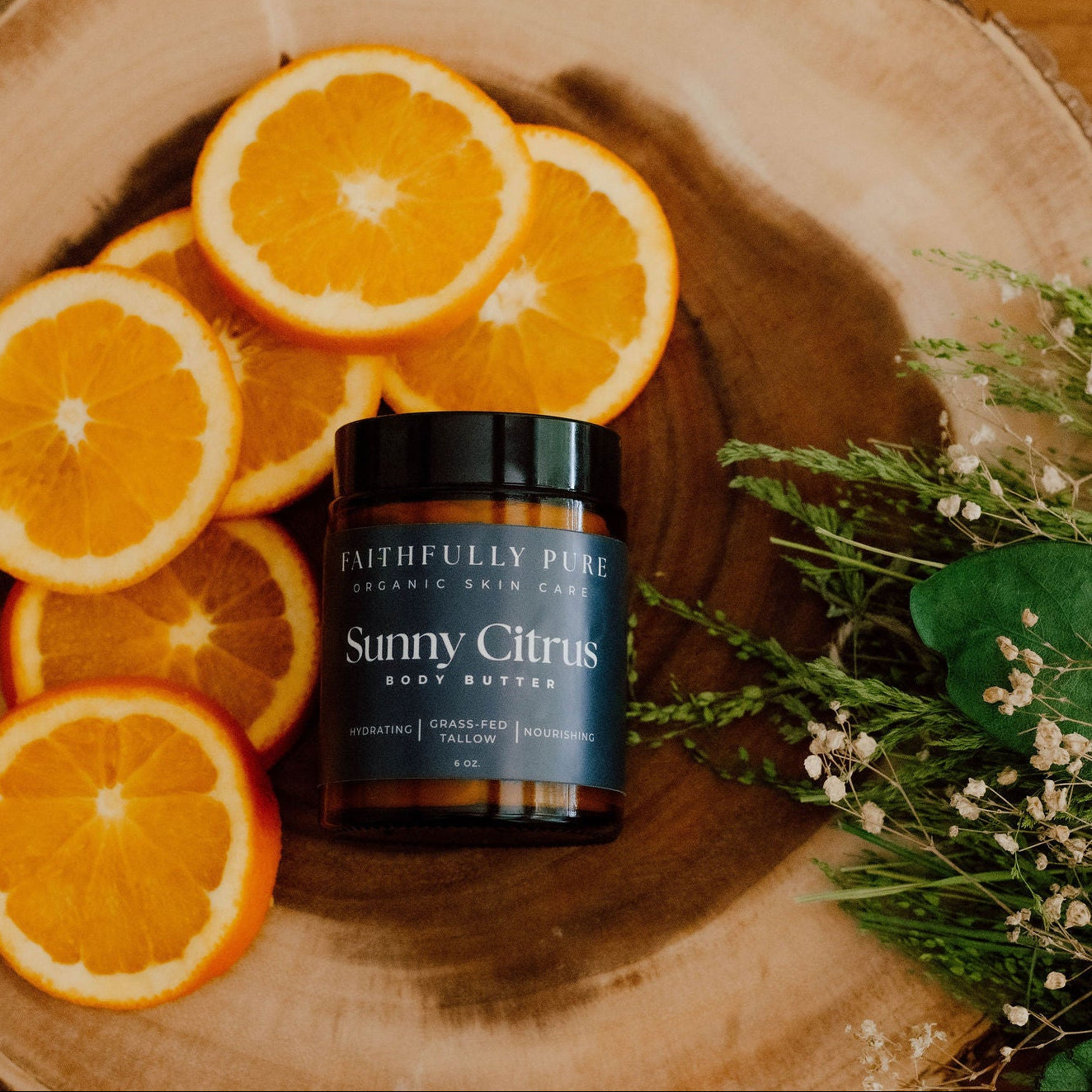 Body butter jar labeled 'Sunny Citrus' surrounded by sliced oranges on a wooden surface.