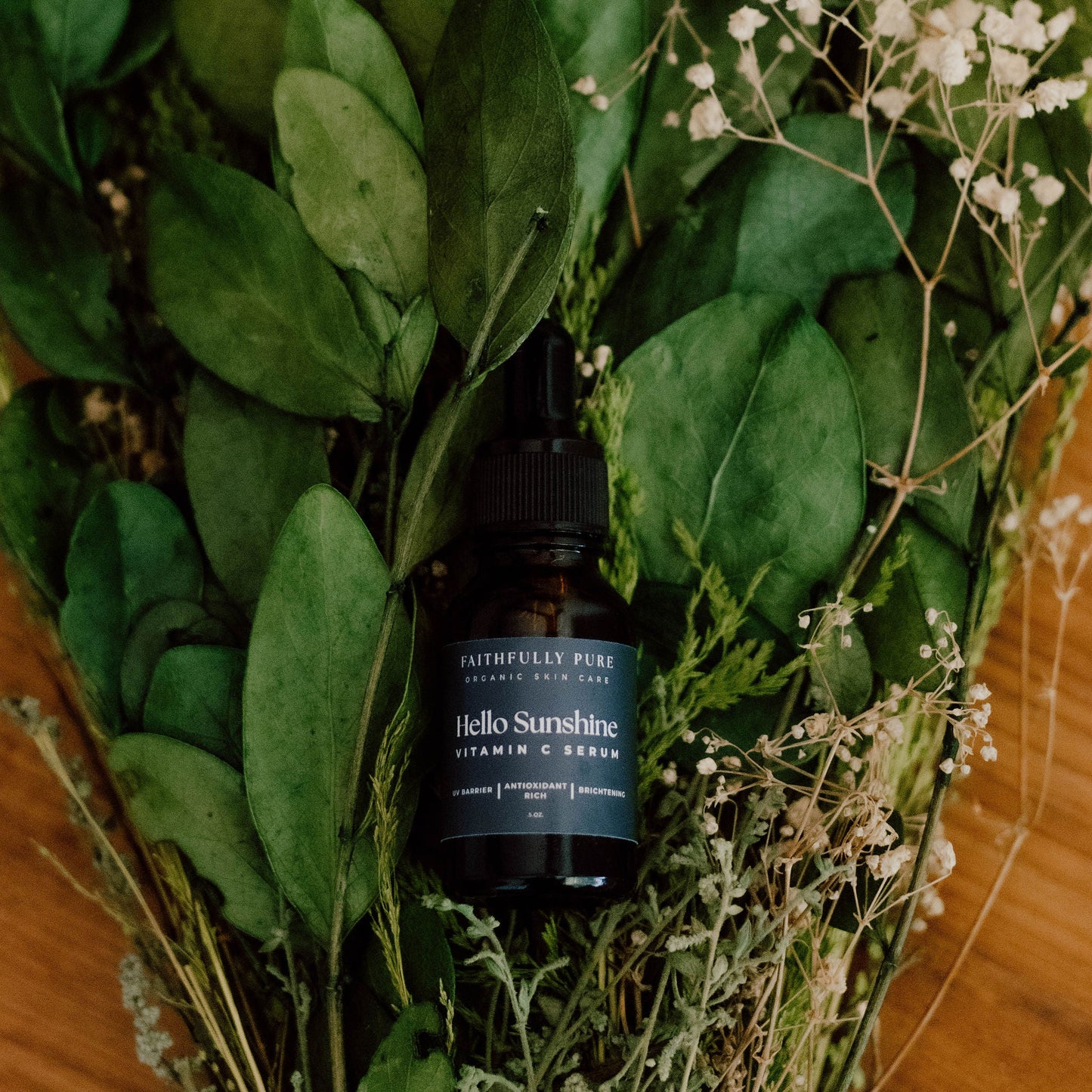 Bottle of Hello Sunshine Vitamin C Serum surrounded by green leaves and small white flowers on a wooden surface